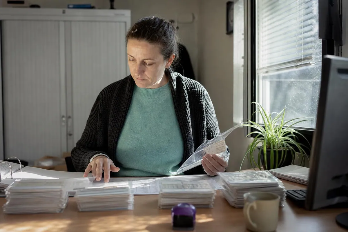 Une trésorière d'amicale du personnel assise à un bureau administratif, comparant une liste nominative imprimée et des pochettes de bons d'achat classées